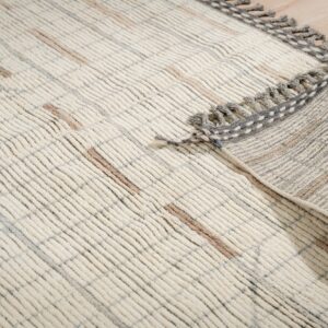 Textured off-white rug with raised ribs and tan accents rests on wood planks, showing a gray fringed border.