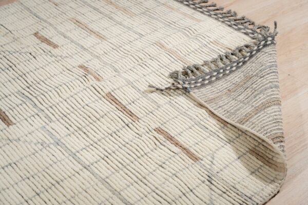 Textured off-white rug with raised ribs and tan accents rests on wood planks, showing a gray fringed border.