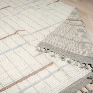 High-pile cream rug with gray and taupe plaid pattern partially rolls back on wood floor, showing tassels.