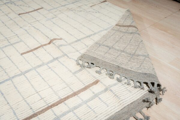 High-pile cream rug with gray and taupe plaid pattern partially rolls back on wood floor, showing tassels.