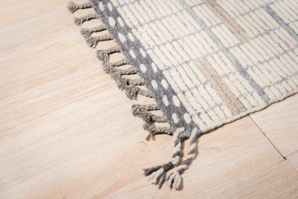 Thick, tufted white rug with a blue and beige checkered pattern and gray knotted fringe rests on light wood flooring.