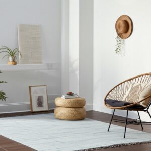 Pale blue area rug with fringe anchors a woven chair and poufs on dark wood flooring.