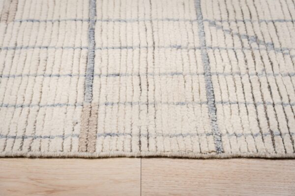 Close-up view of a chunky ivory rug with a sparse blue and tan grid design resting on light wood flooring.