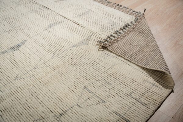 Textured cream rug with gray accents, curled to show the striped woven backing on light wood floor.