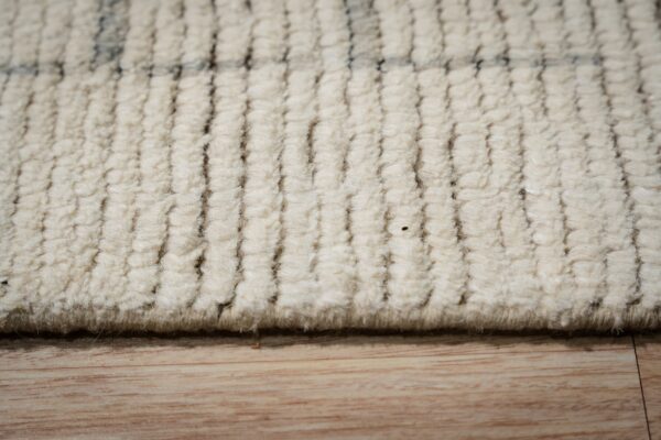 Detailed view of a cream, high-pile textured rug showing ribbed texture and faint dark accents over wood flooring.