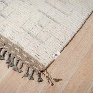 High-pile cream rug with subtle gray patterns, braided tassels, and a woven brown edge on light wood floors.
