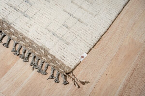 High-pile cream rug with subtle gray patterns, braided tassels, and a woven brown edge on light wood floors.