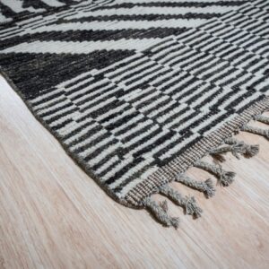 High-pile black and off-white rug with bold geometric patterning and knotted fringe on light wood.