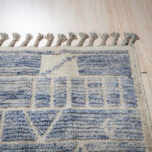Textured beige rug with high-pile blue geometric patterns and fringe on light wood flooring.