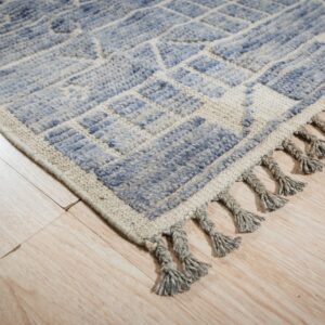 Textured, geometric blue and cream rug edge with gray braided tassels on light wood floor.