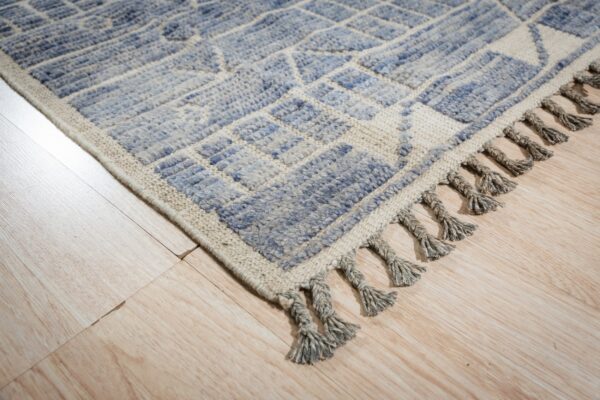 Textured, geometric blue and cream rug edge with gray braided tassels on light wood floor.