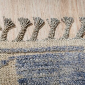 A close-up view of thick, braided tassels on a textured blue and beige rug over light wood flooring.