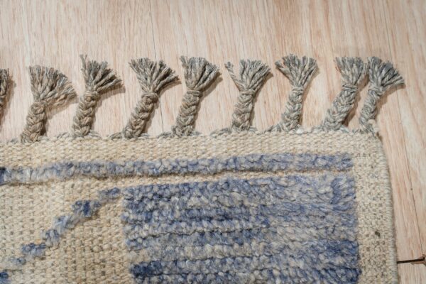 A close-up view of thick, braided tassels on a textured blue and beige rug over light wood flooring.