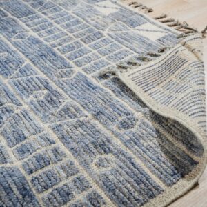 Textured blue and beige geometric rug folded back, showing fringe and striped backing on light wood.