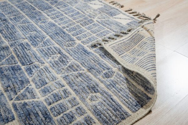 Textured blue and beige geometric rug folded back, showing fringe and striped backing on light wood.