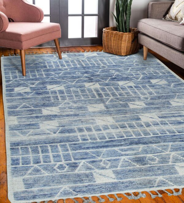 Blue and light geometric rug with tassels on a wood floor next to pink and gray seating.