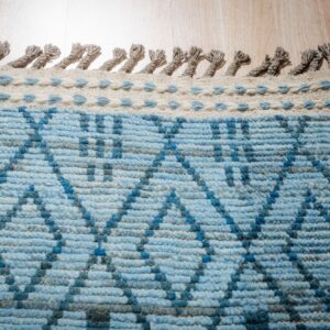 Low pile blue rug with geometric pattern, cream border, and knotted gray fringe on wood floor.