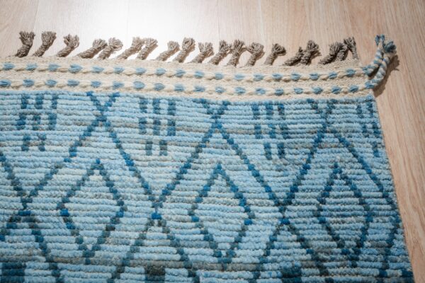 Low pile blue rug with geometric pattern, cream border, and knotted gray fringe on wood floor.
