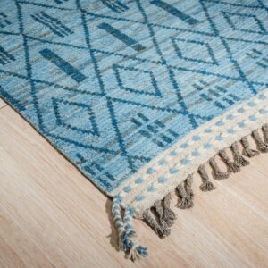 Close-up of a blue and gray patterned rug with diamond designs, light flooring, and braided gray tassels.