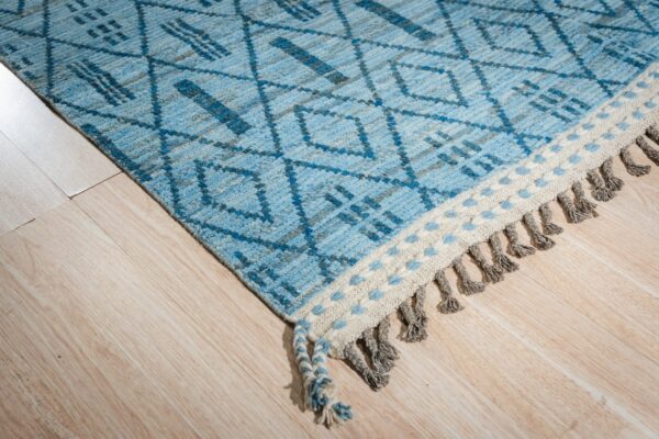 Close-up of a blue and gray patterned rug with diamond designs, light flooring, and braided gray tassels.