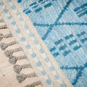 Close-up of a textured blue and cream rug with a geometric pattern and braided gray fringe on wood floor.