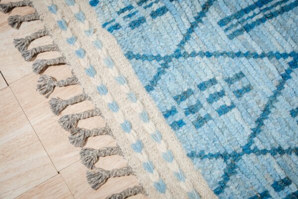 Close-up of a textured blue and cream rug with a geometric pattern and braided gray fringe on wood floor.