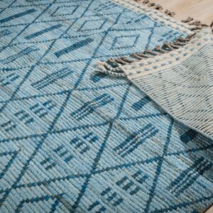 Textured blue pile rug showing a geometric diamond pattern and backing detail with gray fringe on pale wood planks.
