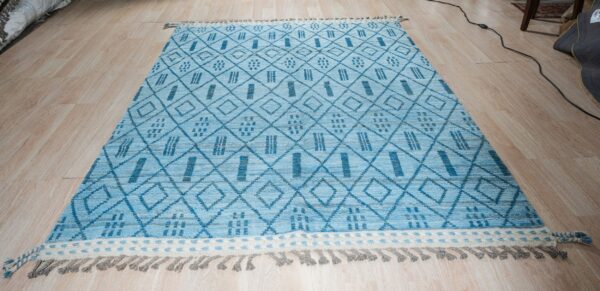 Blue high-pile rug with geometric diamond and stripe motifs, fringed ends, displayed on wood planks.