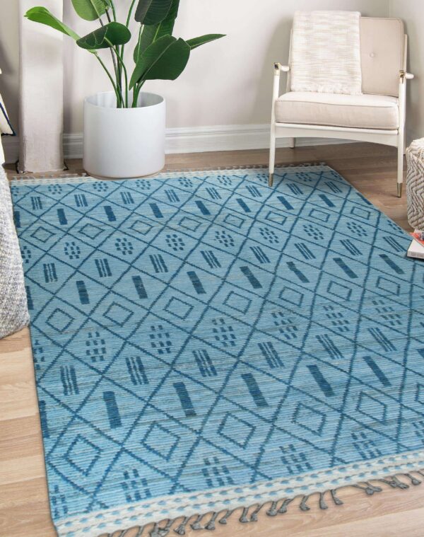 Textured blue flatweave rug with dark diamond patterns lies on hardwood floor near a plant and white chair.