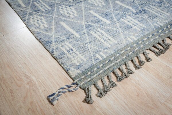 Textured light blue and white geometric rug with braided gray tassels resting on light wood flooring.