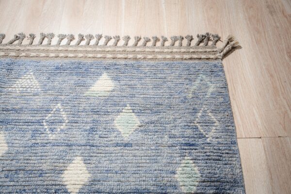 Textured blue rug featuring diamond patterns, a gray border, and braided tassels on light wood planks.