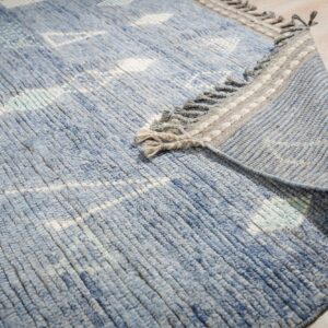 Blue textured rug with white patterns partially folded over, showing a striped backing on light wood.