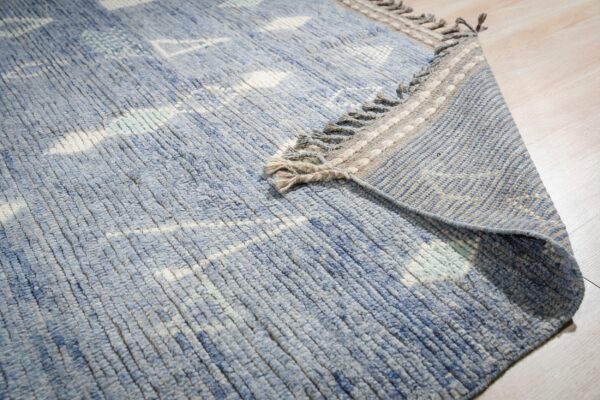 Blue textured rug with white patterns partially folded over, showing a striped backing on light wood.