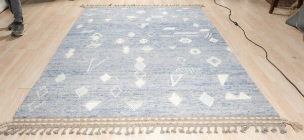 Textured light blue rug with scattered white geometric patterns and gray braided tassels on a light wood floor.