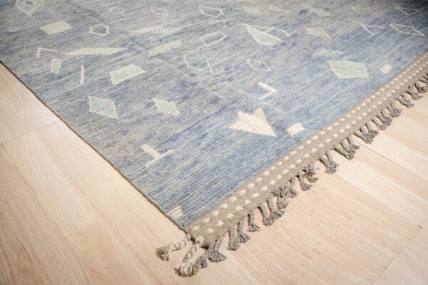 A textured blue rug featuring white geometric designs and a dotted fringe border rests on light wood flooring.