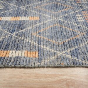 Textured blue rug showing geometric pattern in orange and white on light wood plank flooring.