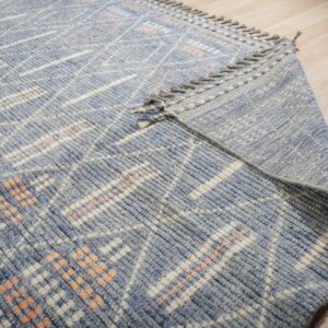 Textured blue rug with white and peach geometric pattern shown folded over on light wood flooring.