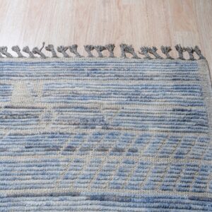 Blue and beige high-low textured rug with thick gray tassels shown on light wood flooring.