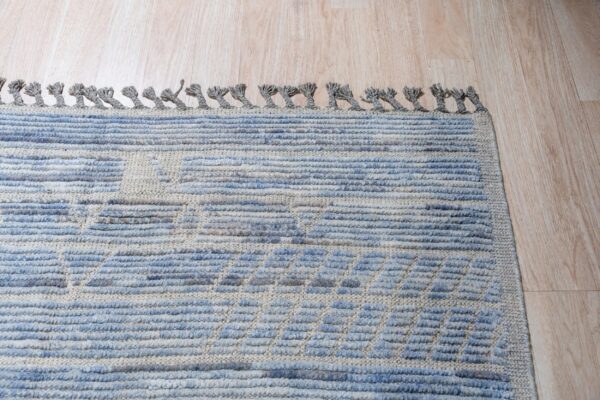 Blue and beige high-low textured rug with thick gray tassels shown on light wood flooring.