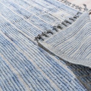 Close-up of a blue and gray textured rug with thick stripes and tassels folded on light wood flooring.