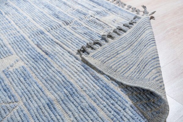 Close-up of a blue and gray textured rug with thick stripes and tassels folded on light wood flooring.