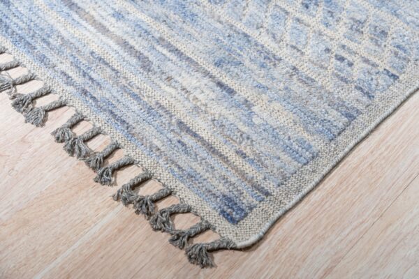Textured blue and gray patterned rug corner with thick tassels rests on light wood planks.