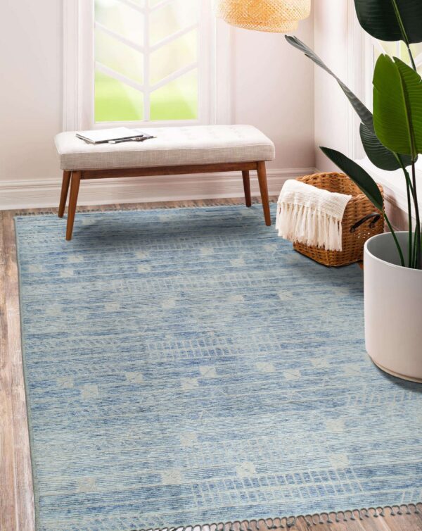 Light blue geometric rug with tassels on hardwood flooring next to a bench and a large houseplant.
