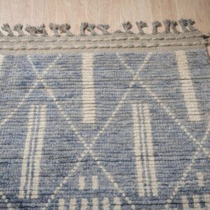 A textured blue and off-white geometric rug border with braided fringe rests on light wood planks.