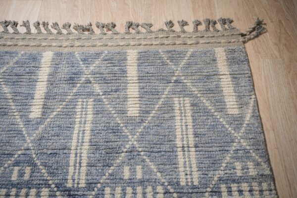 A textured blue and off-white geometric rug border with braided fringe rests on light wood planks.