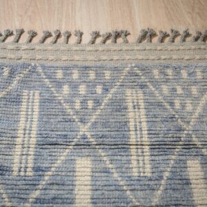 Blue and cream ribbed geometric rug featuring a thick gray knotted fringe along the edge on wood flooring.