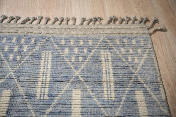 Blue and cream ribbed geometric rug featuring a thick gray knotted fringe along the edge on wood flooring.