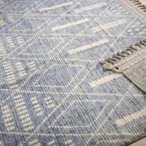 Close-up of a blue and cream textured rug featuring a geometric pattern and gray knotted fringe on wood flooring.