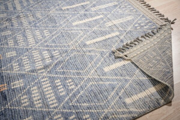 Close-up of a blue and cream textured rug featuring a geometric pattern and gray knotted fringe on wood flooring.
