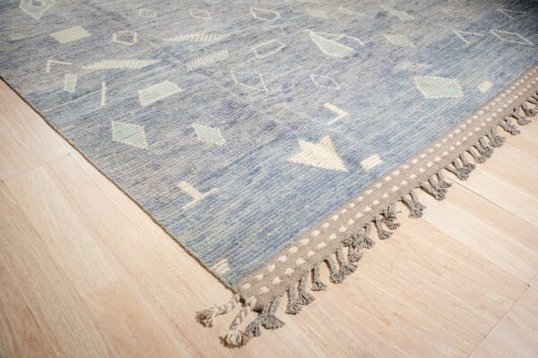 Blue textured rug with white abstract motifs and a gray tasseled border on light wood flooring.